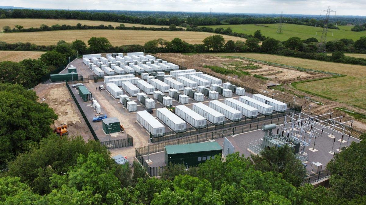 Aerial view of a fenced facility with many white modular containers arranged in rows beside electrical equipment in farmland.