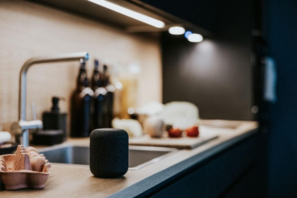 Smart speaker on a kitchen counter beside an egg carton, with blurred bottles and sink in the background.