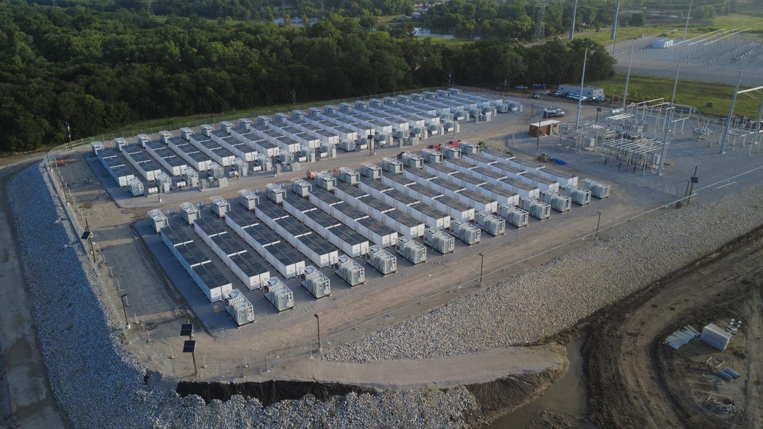 Aerial view of an industrial yard with dozens of white shipping containers arranged in parallel rows, gravel ground, and a rocky shoreline nearby.