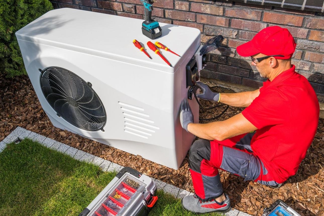 HVAC technician in red kneels beside outdoor AC condenser, using a tool on the access panel with a toolbox nearby.