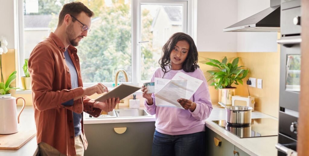 Man and woman stand at a bright kitchen counter, reviewing documents and the tablet together.