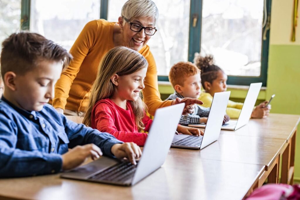 Teacher assists smiling students using laptops at a sunny classroom desk.