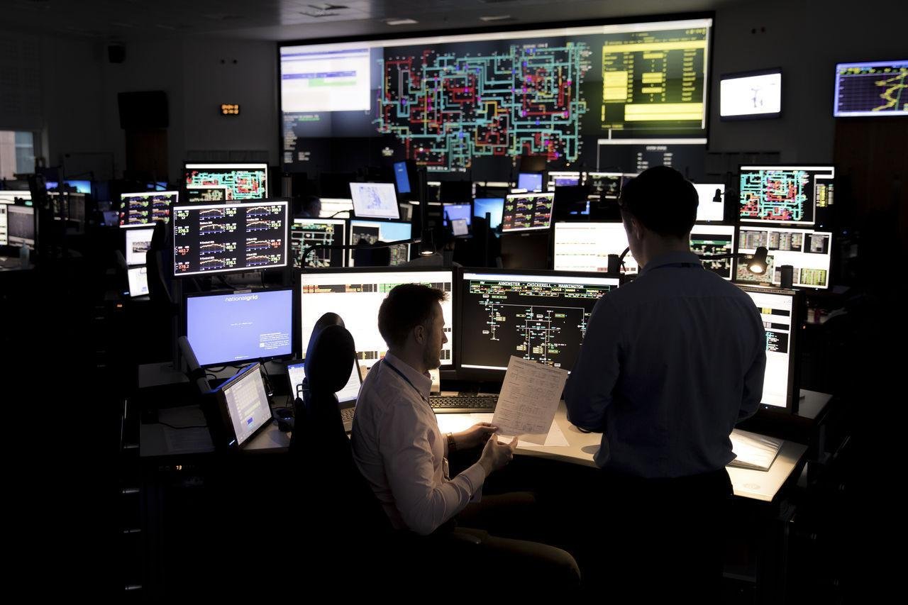 Control room filled with many monitors showing charts and diagrams; two operators review data at a workstation.