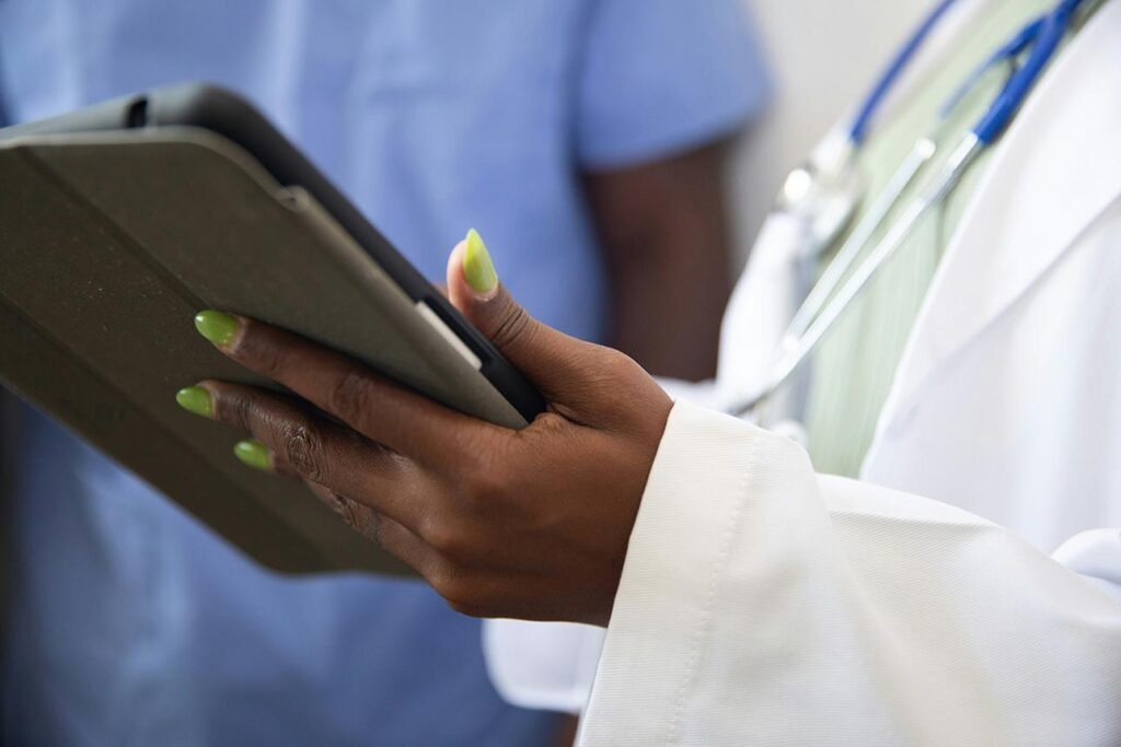 Doctor in a white coat using a tablet to review patient information, with another clinician in blue scrubs in the background.