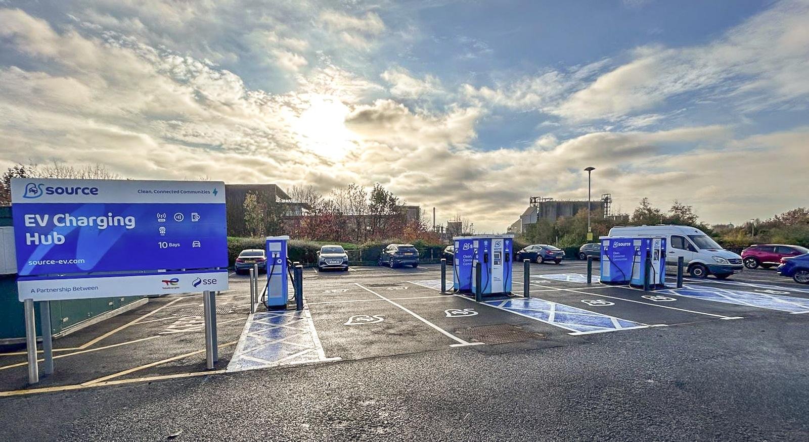 Blue EV charging hub in a parking lot with multiple charging stations and a large sign reading 'EV Charging Hub' and 'source-ev.com' on the left side. Cars and a van parked nearby under a cloudy sky.
