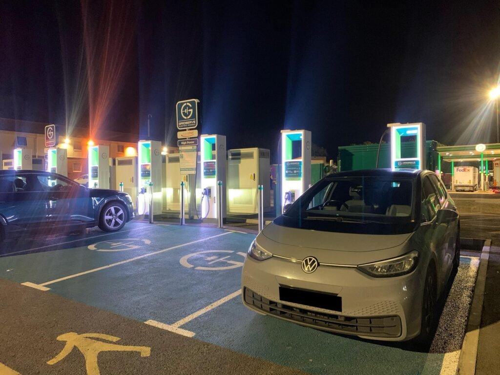 Night scene at an electric vehicle charging station with multiple illuminated charging kiosks and a VW van in the foreground.