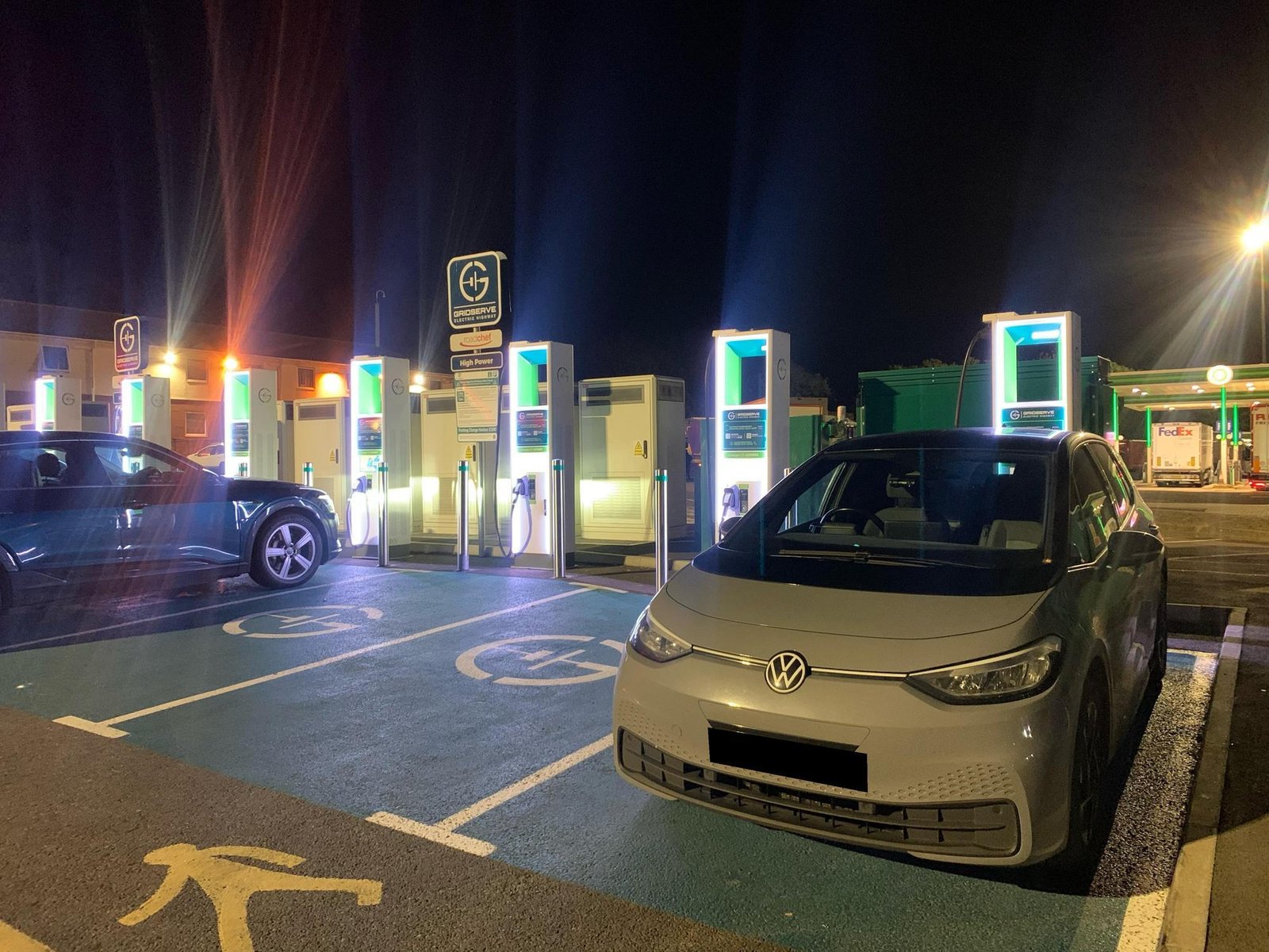 Night scene at an electric vehicle charging station with multiple illuminated charging kiosks and a VW van in the foreground.