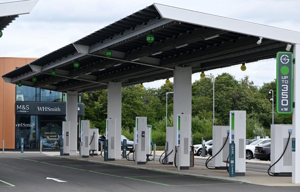 Electric vehicle charging station with multiple white chargers under a large canopy; WHSmith storefront visible in the background.