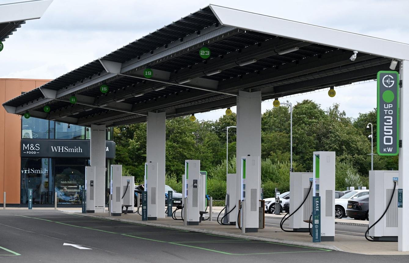 Electric vehicle charging station with multiple white chargers under a large canopy; WHSmith storefront visible in the background.