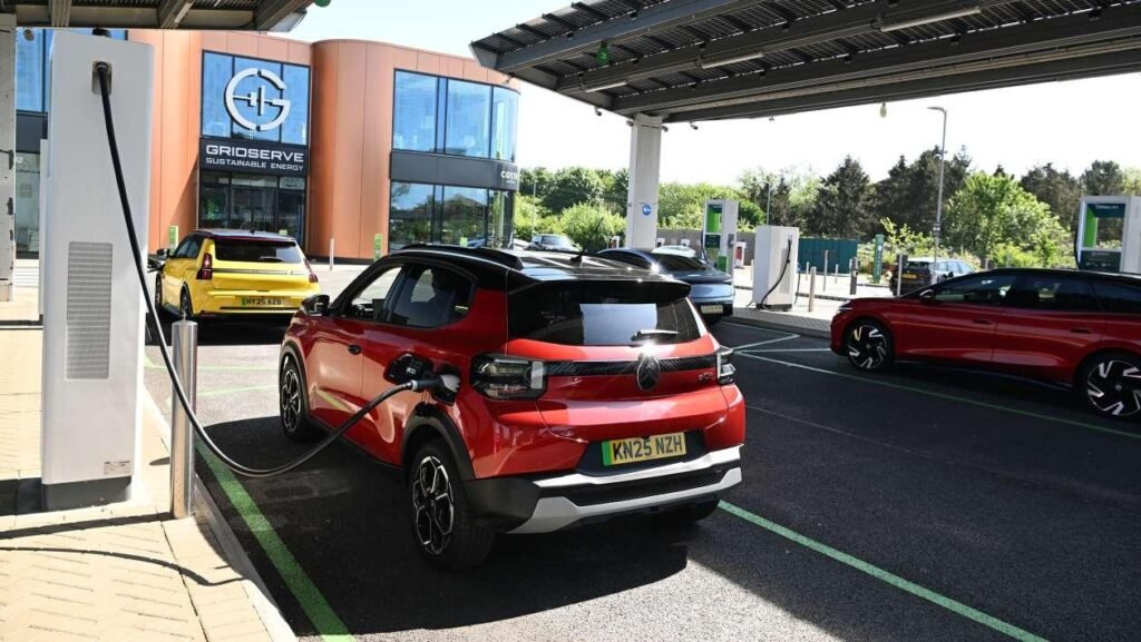 Red electric SUV plugged into a charging station at a Gridserve facility, with another car and modern building in the background.