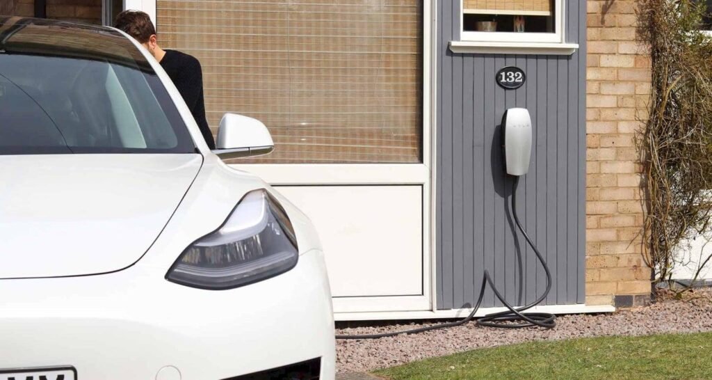 White electric car parked at a home charging station with a wall-mounted charger and charging cable nearby.