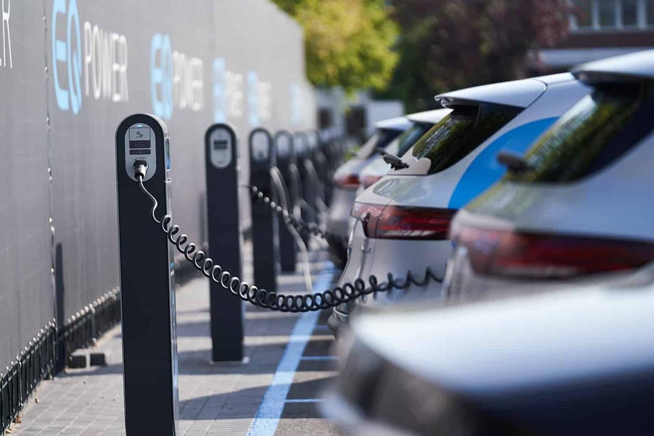 Row of EV charging posts along a parking area with a plugged-in vehicle connected by a coiled cable.
