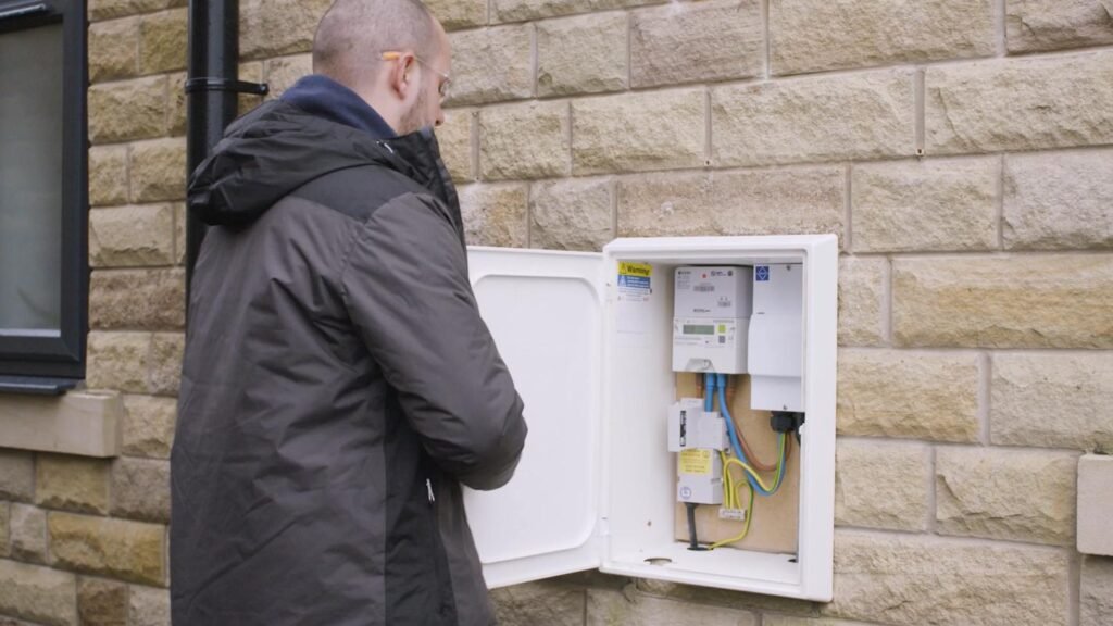 Man in a dark jacket standing by an outdoor electrical meter cabinet mounted on a brick wall, examining the panel door open.