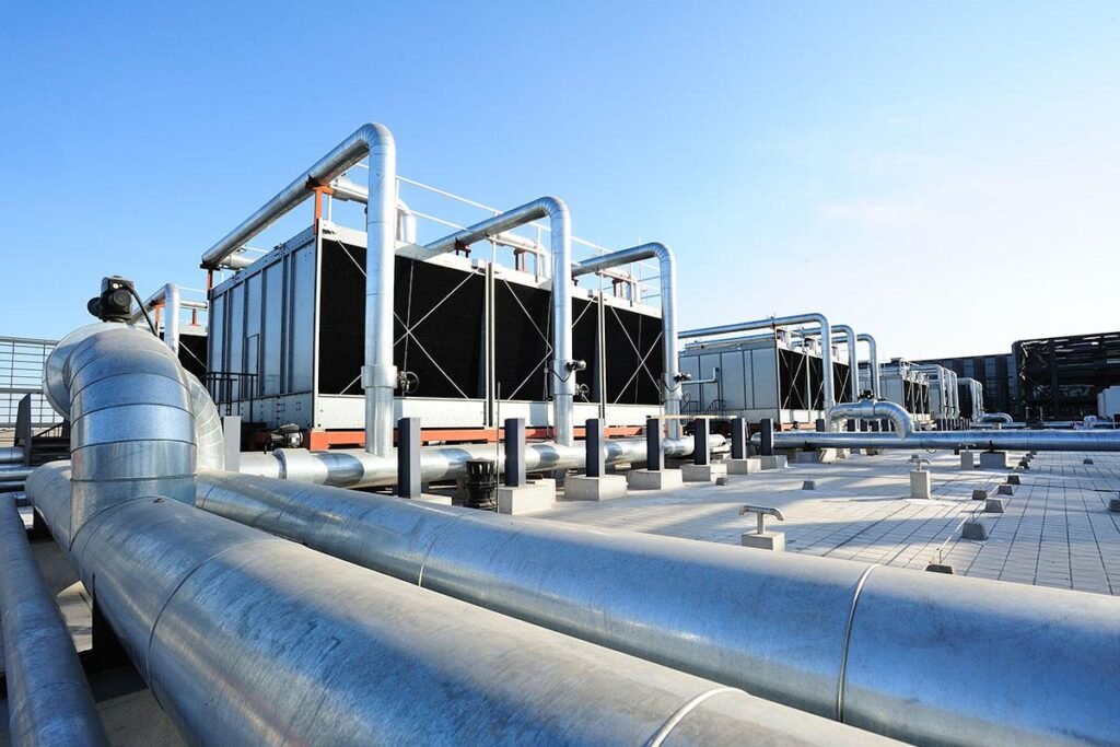 Rooftop view of large industrial cooling units and silver insulated pipes under a clear blue sky.