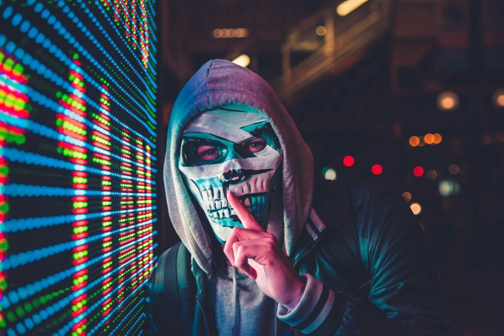 Hacker Shush - Cyber Security England Person wearing a hoodie and skull mask posing beside a wall of colorful LED lights in a dark urban setting.