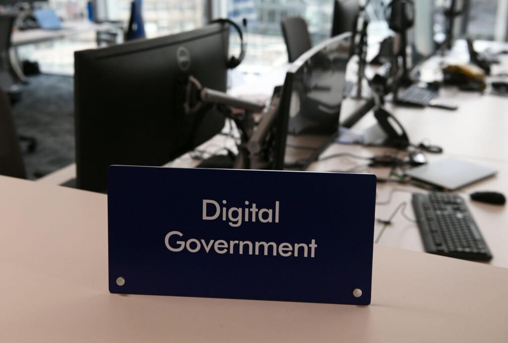 Blue sign reading 'Digital Government' on a desk in an office with computer monitors in the blurred background.