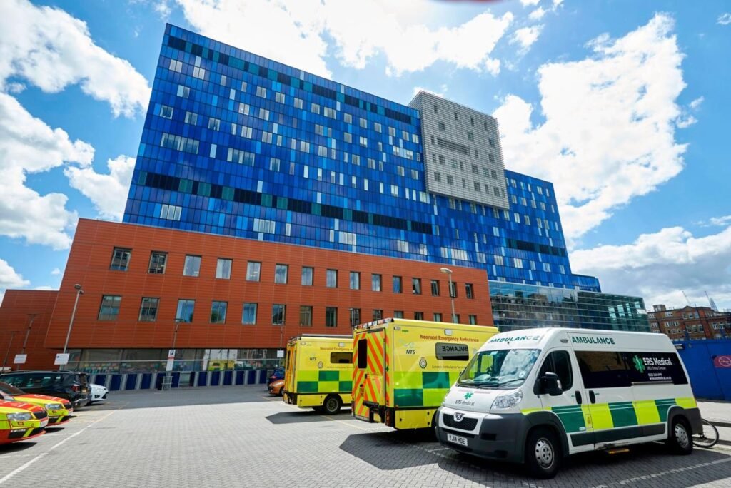 Modern multi-storey blue and orange hospital building with ambulances parked in a paved lot in front.