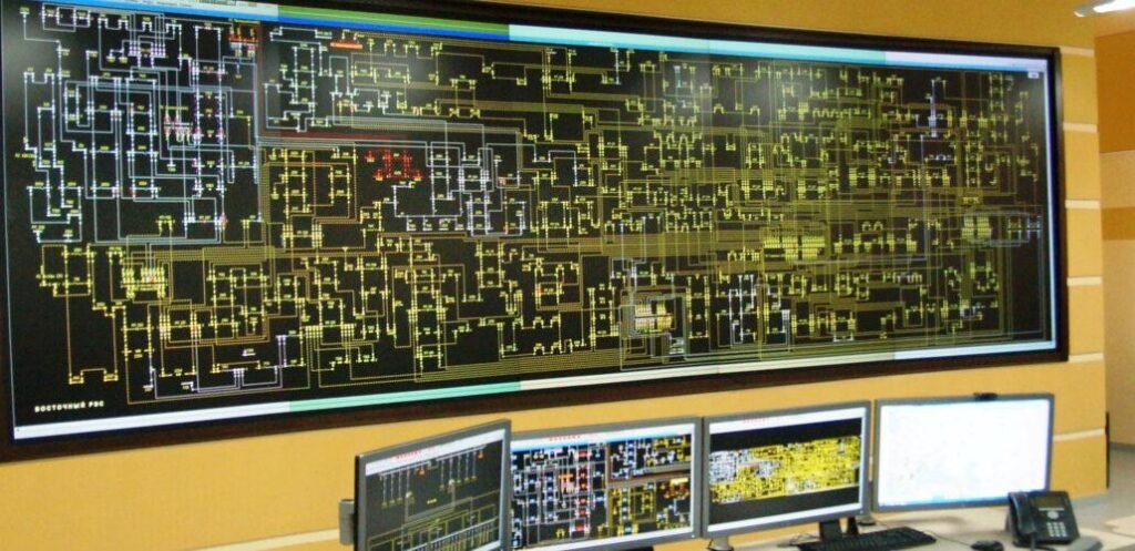 Large wall monitor displaying a dense electronic circuit schematic with interconnected lines and components in a control room, with several desktop screens below.