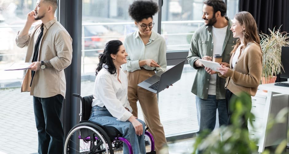 Diverse team in a bright office; a woman in a wheelchair uses a laptop while colleagues chat and share documents.