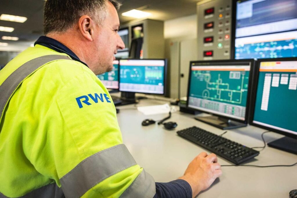 Male technician in a high-visibility yellow jacket sits at a desk with multiple monitors displaying control room dashboards and charts.
