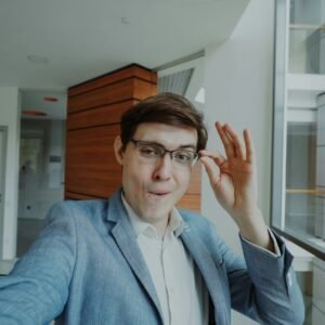 Man in a light blue blazer adjusts his glasses in a bright, modern office lobby with large glass windows behind him.