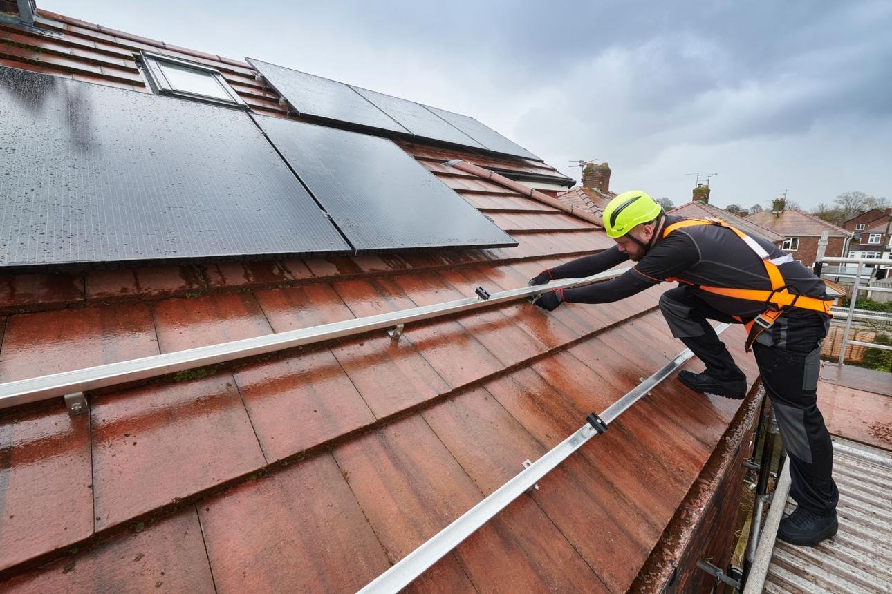 Worker in a yellow helmet and safety harness installs a metal rail along a wet red-tiled roof beside solar panels on a cloudy day.