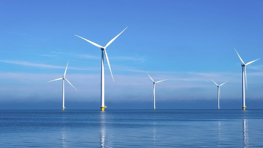 Offshore wind turbines in the calm blue sea with reflections on the water under a clear sky, six white blades visible.