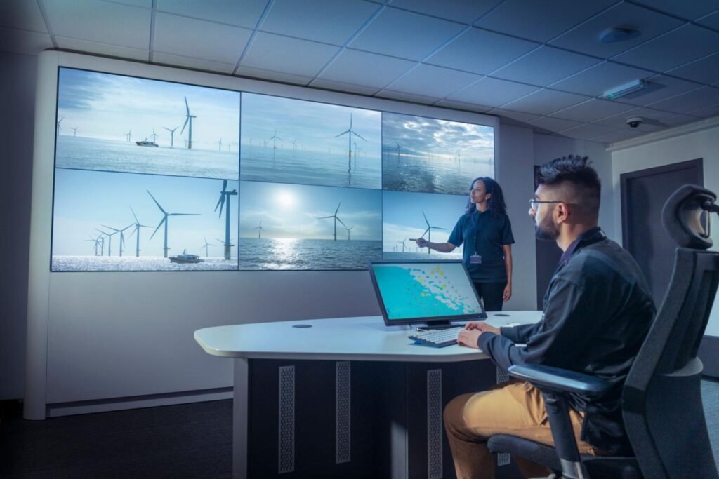 Large video wall showing offshore wind turbines; a presenter points to the screens while a man sits at a desk with a laptop in a modern control room.