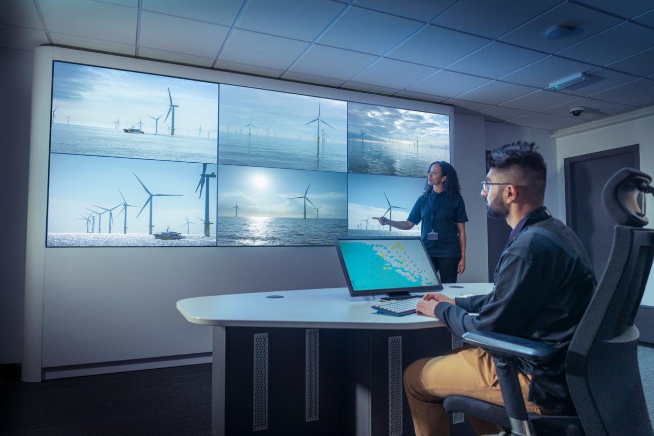 Large video wall showing offshore wind turbines; a presenter points to the screens while a man sits at a desk with a laptop in a modern control room.