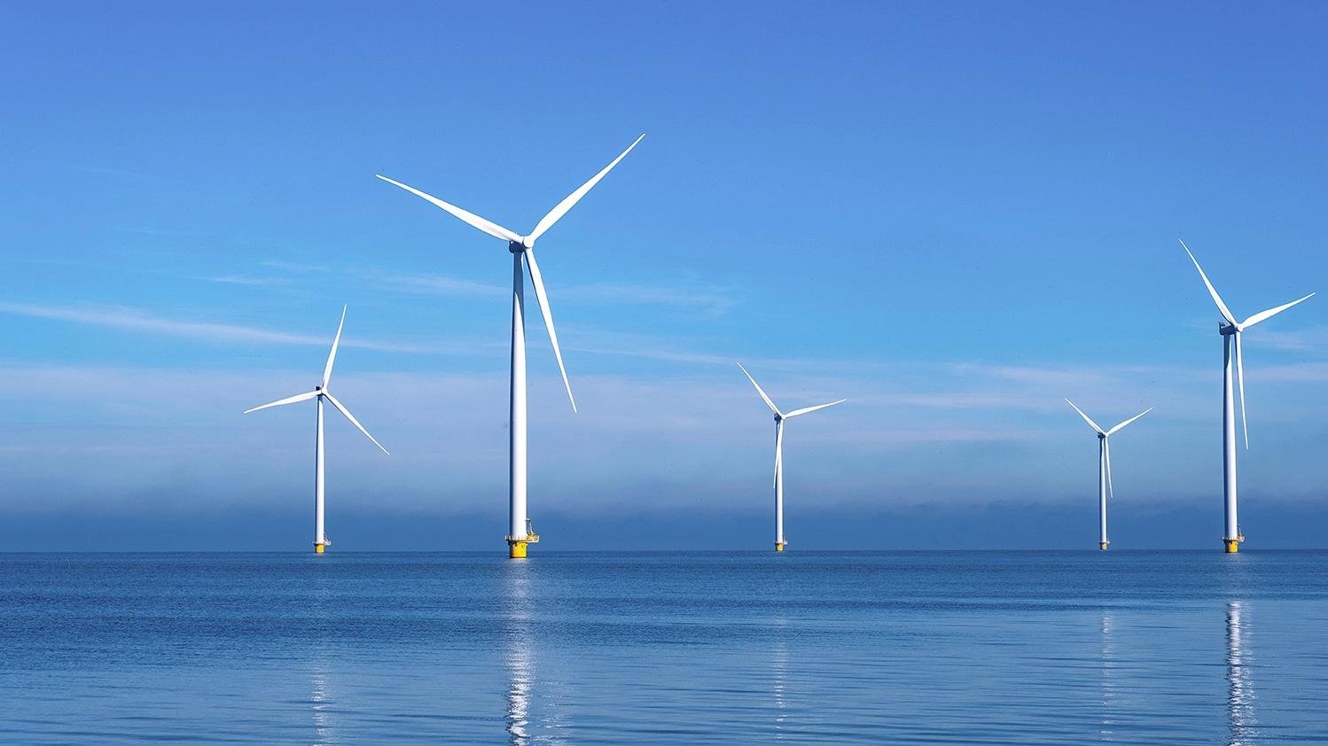 Offshore wind turbines in the calm blue sea with reflections on the water under a clear sky, six white blades visible.