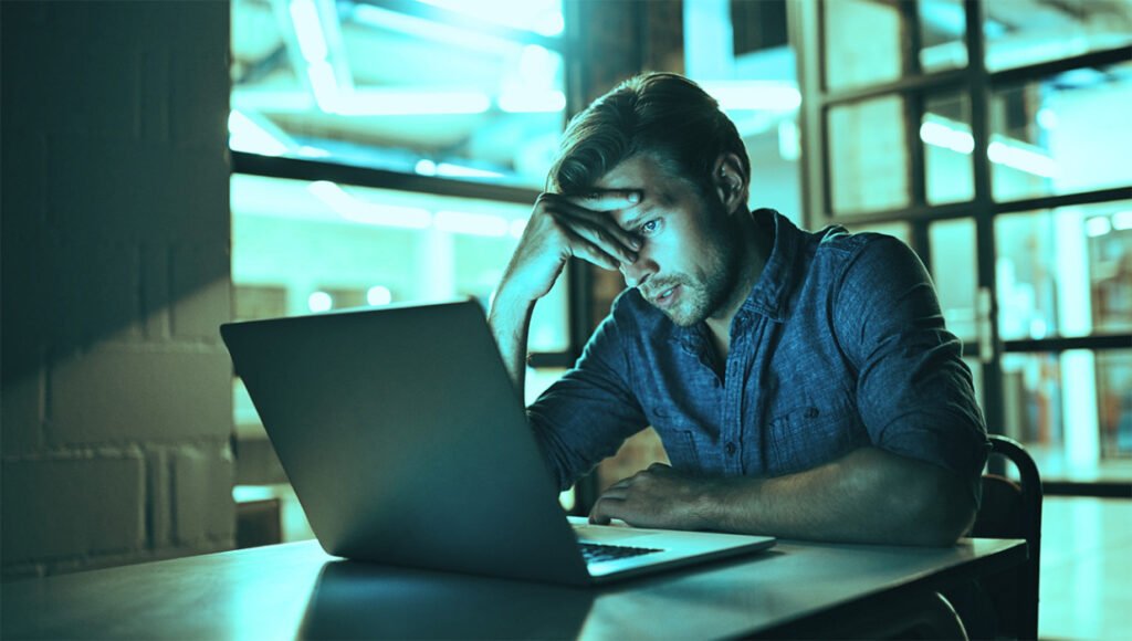 Man sitting at a table working on a laptop, his hand resting on his forehead as he looks at the screen in a neon-lit space.