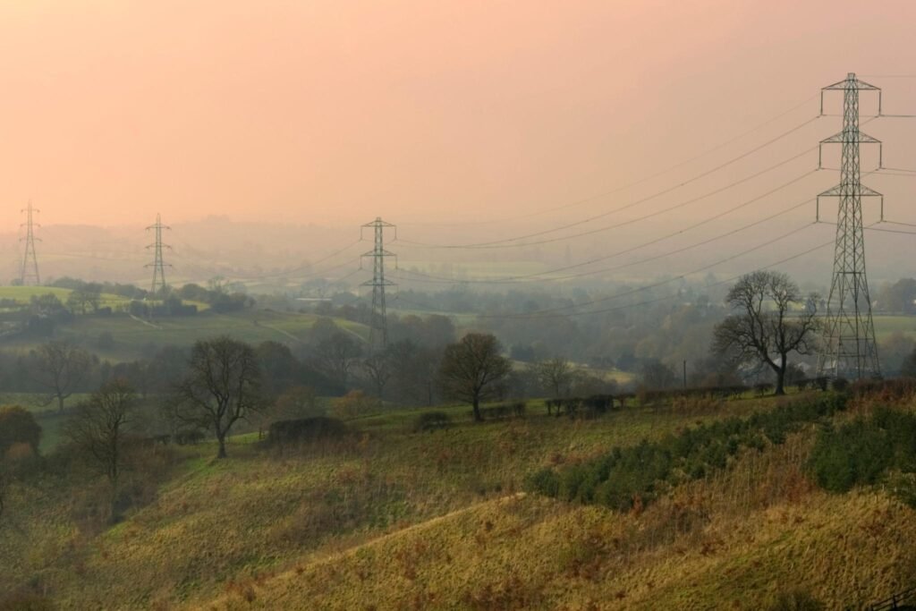 Rural landscape at sunrise with tall electricity pylons and power lines crossing rolling fields and trees.