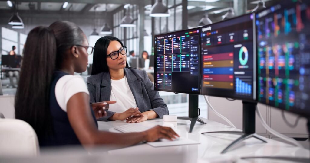 Two women collaborate at a desk, studying dashboards on multiple monitors showing data charts and analytics in a modern office.