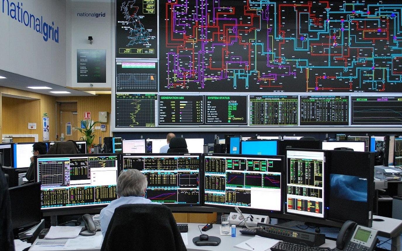 Control room with many computer monitors showing data dashboards and a large wall display of color-coded grid diagrams above the desks.