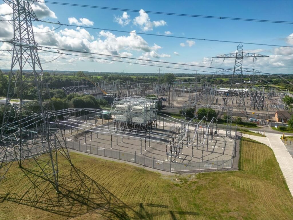 High-voltage substation with metal lattice towers, transformers, and fenced equipment under a blue sky.