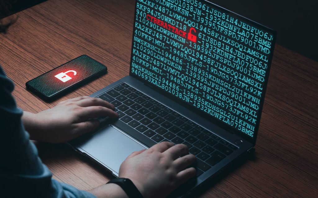 Laptop screen shows blue code with a red padlock, while a smartphone on the desk displays a lock icon; hands type on the keyboard (cybersecurity scene)