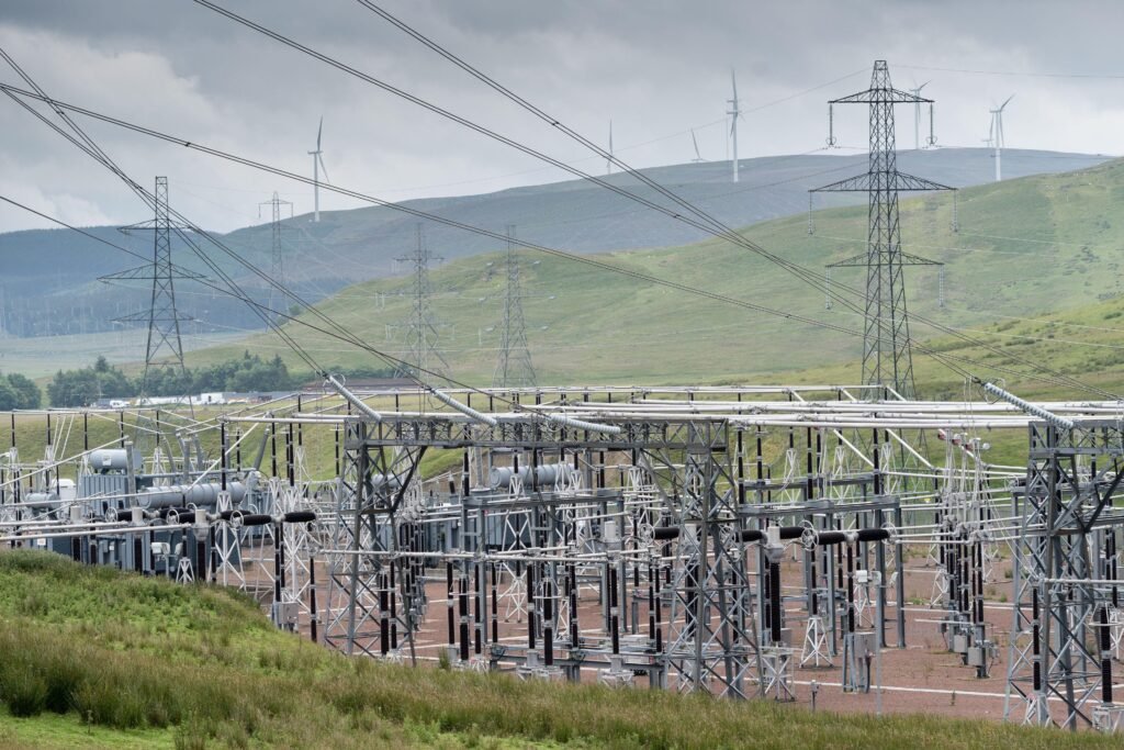 High-voltage transmission towers and overhead cables stretch across a grassy landscape, with wind turbines on distant hills and cloudy sky in the background.