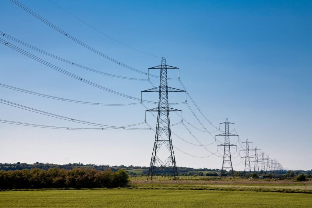 Row of high-voltage transmission towers with wires crossing a blue sky over a rural landscape.