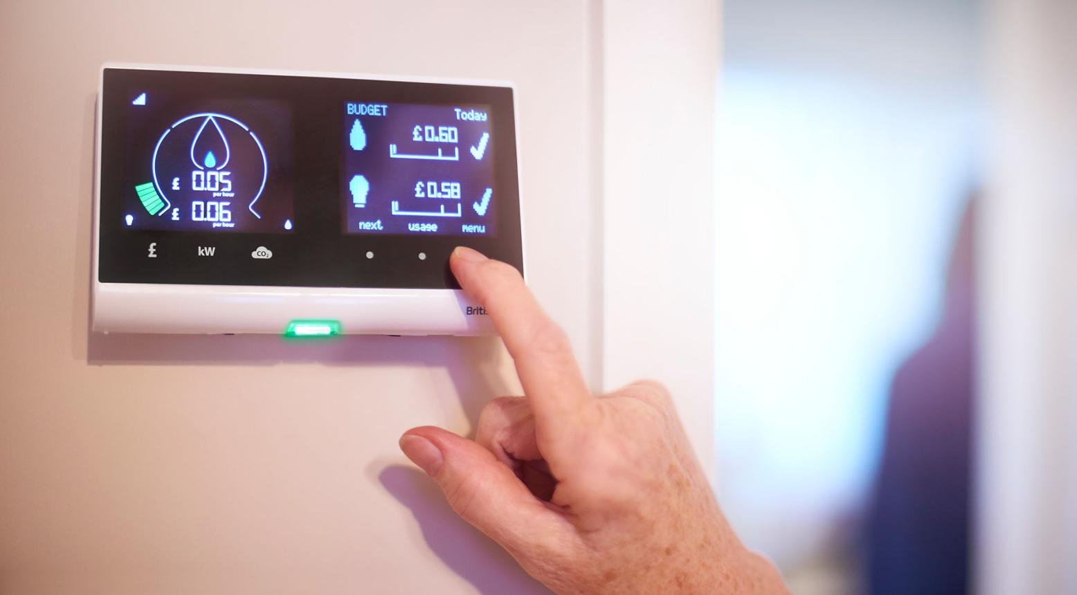 Hand adjusts a wall-mounted digital energy monitor showing budget and usage data on a blue-lit screen.